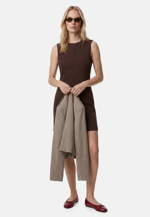 Donna con abito marrone scuro senza maniche, che tiene una giacca beige a righe, indossa occhiali da sole e ballerine rosse, in piedi davanti a uno sfondo bianco.