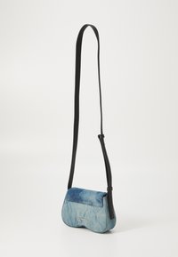 Džínová crossbody taška se zakřiveným tvarem, modré barvy, černým koženým popruhem a stříbrným logem vpředu.