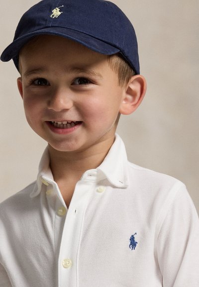 Joven niño sonriente, con una gorra azul marino y una camiseta polo blanca con un logo de caballo bordado en azul.