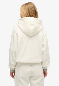 Mujer con cabello rubio que lleva una sudadera crema y pantalones a juego, de pie de espaldas a la cámara contra un fondo simple.