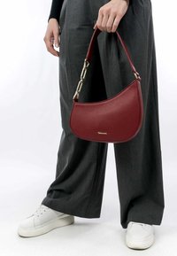 Borsa a mano in pelle rossa a ?righe, con forma arrotondata, dettagli in metallo dorato e manico corto, tenuta da una persona che indossa pantaloni larghi grigi e sneaker bianche.