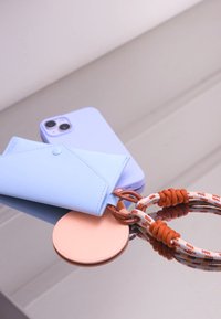 Portacarte in pelle azzurro chiaro, smartphone azzurro pastello, etichetta rotonda in pelle color pesca e corda intrecciata con accenti ruggine e bianchi su una superficie riflettente.