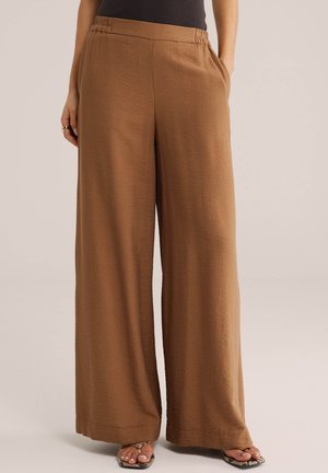 Žena oblečená v hnedých nohaviciach s vysokým pásom a širokými nohavicami so zapustenými bočnými vreckami a čiernom topu, stojaca s rukami vreckách na jednoduchom pozadí.