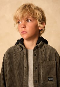 Veste-chemise en velours côtelé vert olive avec des boutons-pression noirs et une poche avant, portée par-dessus un sweat-shirt gris, doté d'une capuche.