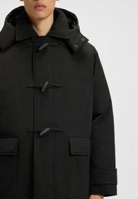 Parka nera imbottita con chiusure a cordoncino, tasche con pattina, colletto alto e cappuccio staccabile con bottone a pressione su una parte superiore del corpo di una persona.