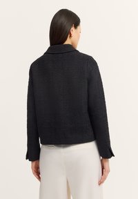 Schwarze texturierte Jacke mit Kragen, kurzem Design, langen Ärmeln und dezenten Schlitzen an den Manschetten, von hinten präsentiert.