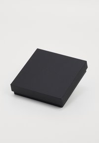 Melnas kvadrātveida kaste ar nedaudz teksturētu virsmu un smalku reljefa zīmolu uz vāka, novietota uz baltas fona.