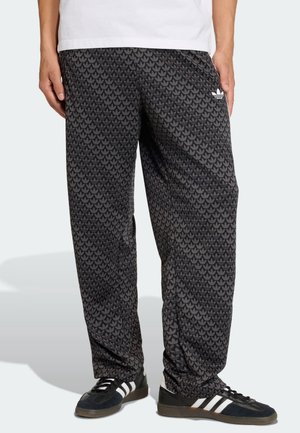 Homme portant un pantalon de survêtement Adidas noir à motifs et des baskets Adidas noires avec des bandes blanches, debout devant un fond clair.