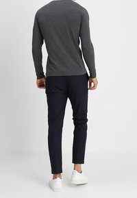 Mann trägt ein dunkelgraues Langarmshirt, marineblaue slim-fit Hosen und weiße Sneaker und steht vor einem einfarbigen hellen Hintergrund.