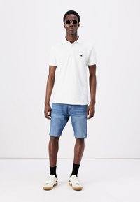 Fehér póló, kicsi logóval, kék farmernadrággal, fekete zoknikkal és fehér sneakerrel párosítva. A modellen kerek, sötét napszemüveg van.