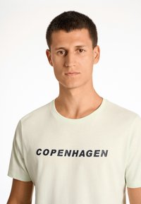 Camiseta de manga corta verde claro con cuello redondo, que presenta la palabra "COPENHAGUE" en texto negro y naranja en negrita en la parte frontal.