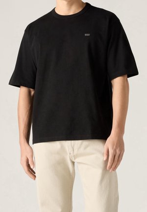 Mand iført en ensfarvet sort kortærmet T-shirt med et lille logo på brystet og lyse beige bukser mod en neutral baggrund.