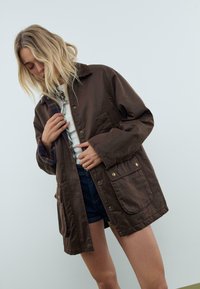 En blond kvinde iført en mørkebrun lynlåset frakke over en hvid top og mørke shorts, justerer sin jakke, mens hun ser ned mod en ensfarvet baggrund.