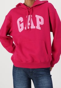 Osoba v jasně růžovém mikině GAP s přední kapsou a šňůrkami, v kombinaci s tmavě modrými džíny na jednobarevném pozadí.