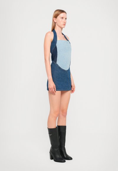 Denim halter obleka z mešanico svetlih in temnih modrih panelov, prilegajoča silhueta in gladka tekstura tkanine. V kombinaciji z črnimi čevlji do kolen.