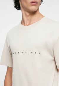 Beige bomulds t-shirt med rund hals og korte ærmer, prydet med et sort "JACK & JONES" logo centreret på fronten.