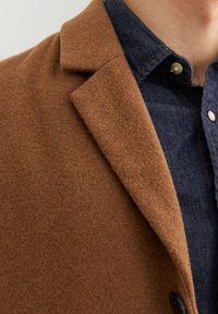 Cappotto di lana marrone con revers a punta e un bottone nero, abbinato a una camicia di denim scuro con colletto a bottoni.