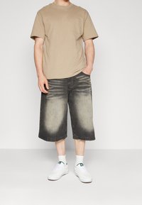 Camiseta de algodón beige con bolsillo, combinada con pantalones cortos de mezclilla negros hasta la rodilla con una textura desgastada. Usada con zapatillas blancas y calcetines.