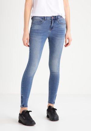 Persoon draagt lichtblauwe skinny jeans met enkelritsen en zwarte sneakers, staand tegen een witte achtergrond.
