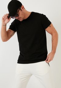 Maglietta nera in cotone con collo rotondo, abbinata a pantaloni bianchi slim-fit. Il modello indossa occhiali da sole e un cappellino nero.