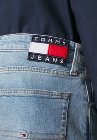 Jeans de mezclilla azul claro con un bolsillo trasero y un parche rectangular del logo de Tommy Jeans en blanco, rojo y azul marino en la cintura, combinados con una camiseta de un tono azul marino oscuro.