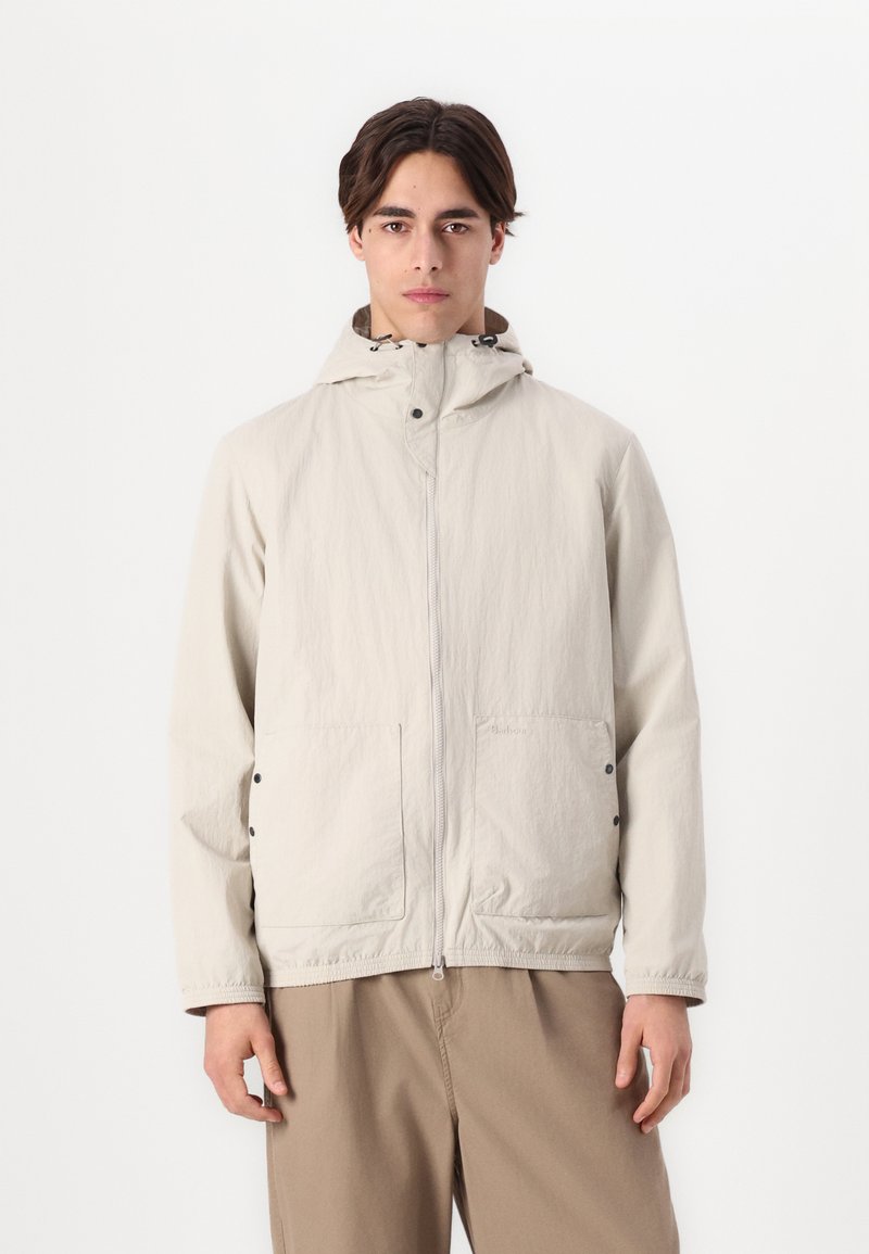 Lichte beige jas met een capuchon, een ritssluiting aan de voorkant en twee zijzakken. Lichtjes gestructureerde stof en elastische manchetten. Subtiel merkdetail.