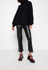 Persoon in een zwart coltrui, zwarte leren cropped broek en donderrode chunky plateau schoenen, staande op een witte vloer.