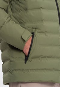 Olivově zelená péřová bunda se zipovou kapsou, mírně prošívanou texturou a nastavitelným manžetem. Obsahuje černý zip a detaily zapínání.