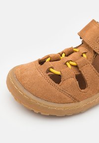 Brune semskede sandal med gule snørebånd, udskårne sider og en holdbar gummisål. Har en velcrostrop til nem justering.
