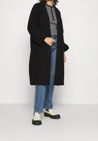 Persona que lleva un largo abrigo negro sobre una camisa de botón a rayas, unos vaqueros azules y botas blancas hasta el tobillo con suelas negras.