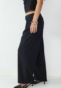 Czarne szerokie spodnie w teksturowanej tkaninie, z wysokim stanem i luźnym krojem, w połączeniu z czarnymi spiczastymi szpilkami.