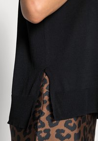 Zbliżenie osoby noszącej czarny dziany sweter z bocznym rozcięciem, nałożony na brązowe i czarne spodnie w panterkę.