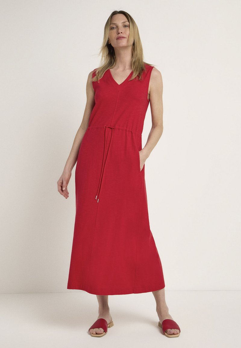 Rotes, ärmelloses Maxikleid mit V-Ausschnitt, Bindegürtel in der Taille und seitlichen Taschen. Glattes Material, knöchellang, kombiniert mit roten Rutschschuhen.