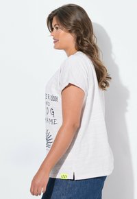 Weißes T-Shirt mit kurzen Ärmeln und horizontalen Streifen, ausgestattet mit einem grafischen Druck in Schwarz und einem kleinen neon-gelben Etikett am Saum.