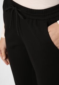 Pantalones negros de cintura elástica con cordón, que presentan bolsillos laterales y una textura suave. Primer plano de la cintura y mano colocada en la cadera.