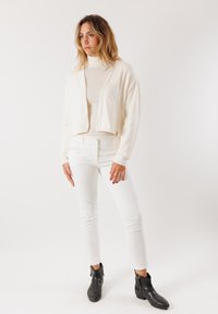 Cardigan bianco corto su un maglione leggero a collo alto, abbinato a pantaloni bianchi aderenti e stivali neri alla caviglia. Texture morbide, design minimalista.