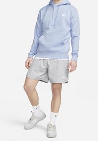 Ljusblå hoodie med en framficka och vit logotyp, ihop med ljusgrå shorts med vit logotyp, vita strumpor och grå sneakers.