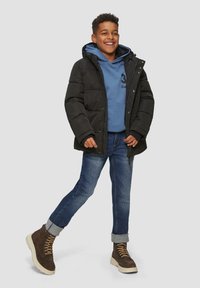 Veste en duvet noire, sweat à capuche bleu en dessous, jeans en denim, bottes marron. Vue en plein corps d'un enfant souriant sur un fond neutre.