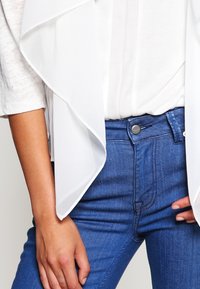 White chiffon overlay drapes over a light top, paired with medium-blue denim jeans featuring a button closure and front pockets.