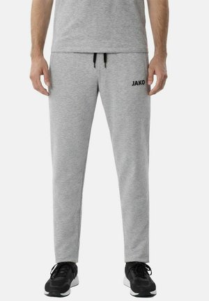 Mann trägt hellgraue JAKO-Sweatpants und passendes Shirt, kombiniert mit schwarzen Sportschuhen, stehend vor weißem Hintergrund.