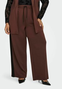 Žena nosi smeđe široke hlače s crnim stranama, crnu čipkastu bluzu dugih rukava i crne cipele s visokom potpeticom s oštrim vrhom.