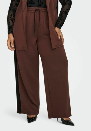 Kvinde iført brune bukser med brede ben og sorte sidestriber, sort blonde top med lange ærmer og sorte spidse højhælede sko.