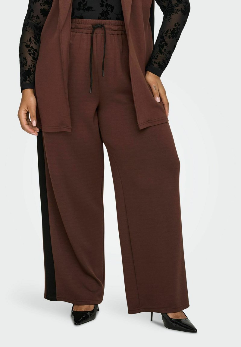 Žena nosi smeđe široke hlače s crnim stranama, crnu čipkastu bluzu dugih rukava i crne cipele s visokom potpeticom s oštrim vrhom.