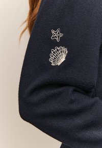 Mörkblå tröja med ärm prydd av en vit broderad sjöstjärna och snäcka nära armbågen, med rött hår synligt på sidan.