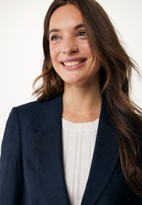 Marinblå blazer med strukturerade slag och långa ärmar, i kombination med en vit texturerad blus med vertikala ränder och scallopad halsringning.