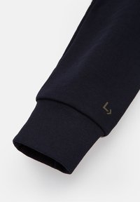 Morskie niebieskie rękawy bluzy w prążkowanej teksturze, z logo w oliwkowej zieleni. Rękaw jest lekko zawinięty na krawędzi.