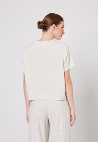 Žena s hnědými vlasy staženými do drdolu, oblečená v volném, světle šedém tričku s krátkým rukávem a odpovídajících kalhotách, stojící zády k divákovi na jednobarevném pozadí.