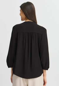 Blusa nera con un motivo a check testurizzato, caratterizzata da una vestibilità rilassata, schiena arricciata e maniche a puff 3/4. L'orlo è leggermente curvo.