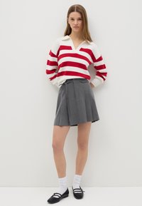 Rot und weiß gestreifter Pullover mit V-Ausschnitt und weiten Ärmeln, kombiniert mit einem grauen, plissierten Rock. Schwarze Schuhe und weiße Socken vervollständigen das Outfit.