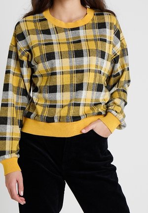 Femme portant un pull à manches longues à carreaux jaune, noir et blanc avec poignets et ourlet côtelés, associé à un pantalon noir, main dans la poche.